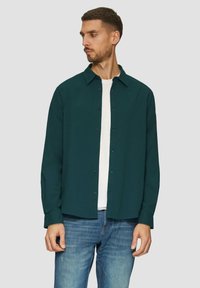 Cyan knoopshirt met een puntkraag, gedragen over een wit t-shirt. De stof heeft een subtiele textuur, gecombineerd met lichtblauwe jeans.