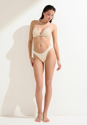 Kobieta stoi przy białej ścianie, ubrana w kremowy szydełkowany top bikini i dopasowane majtki bikini z wysokim stanem, patrzy w dół.