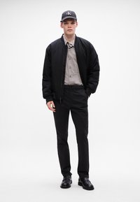 Chaqueta bomber negra sobre una camisa de botones gris, combinada con pantalones negros y zapatos negros brillantes. Gorra negra con el logo "CK" en la parte delantera.