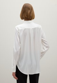 La camisa blanca de manga larga presenta una textura suave y sedosa, un cuello clásico, un detalle de pliegue en la parte posterior y un dobladillo redondeado.