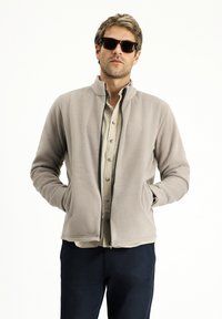 Hellgraue Fleecejacke mit Stehkragen, Reißverschluss und Seitentaschen. Getragen über einem beigen Hemd mit Knöpfen und dunklen Hosen.