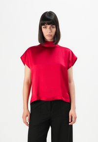 Une femme aux cheveux raides et noirs porte un haut en satin rouge vif à manches courtes et un pantalon large noir devant un fond blanc uni.