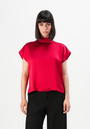 Une femme aux cheveux raides et noirs porte un haut en satin rouge vif à manches courtes et un pantalon large noir devant un fond blanc uni.