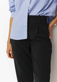 Personne portant une chemise boutonnée à rayures bleu clair rentrée dans un pantalon noir taille haute, avec une main dans la poche et l'autre détendue.