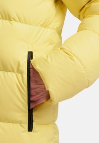 Geel pufferjack met een textuurstof, voorzien van een zichtbare zwarte ritszak aan de zijkant. De mouw heeft een iets puffy design.