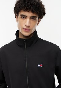 Joven con cabello rizado que lleva una chaqueta negra con cremallera y un pequeño logotipo de Tommy Jeans en rojo, blanco y azul en el pecho.