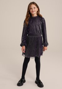 Lila plisserad klänning med långa ärmar, elastisk midja och lager-på-lager kjol. Bärs med svarta strumpbyxor och klumpiga svarta skor. Slät, blank textur.