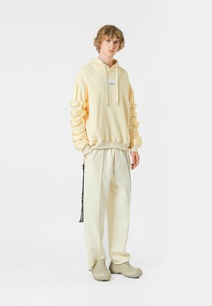 Sudadera amarilla con mangas de textura en capas, combinada con pantalones sueltos de color beige claro y calzado gris texturizado. Detalle de logo simple en la sudadera.