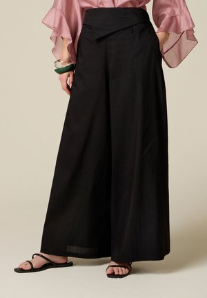 Vrouw draagt zwarte broek met wijde pijpen, doorzichtige roze blouse, zwarte sandalen met bandjes en doorzichtige en groene armbanden aan haar linker pols.