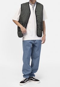 Persona vestida con una camiseta blanca, un chaleco acolchado verde oliva, unos vaqueros azules holgados, unos zapatos deportivos en blanco y negro, y una bufanda de cuello estampada, de pie.