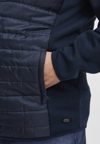 Tamsiai mėlynas džemperis turi siuvinėtą, paminkštintą priekį ir šoninę kišenę. Minkšta medžiagos tekstūra su mažo logotipo žyma apatiniame šone.