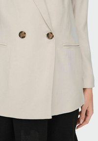 Lichtbeige dubbelzijdig blazer met twee decoratieve bruine knopen, notch revers en zijzakken; glad, op maat gemaakt stof.