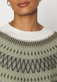 Gestrickter Pullover mit rundem weißem Kragen, gemustert in gedämpften Grün-, Braun- und Creme-Tönen, mit strukturierten geometrischen Designs.