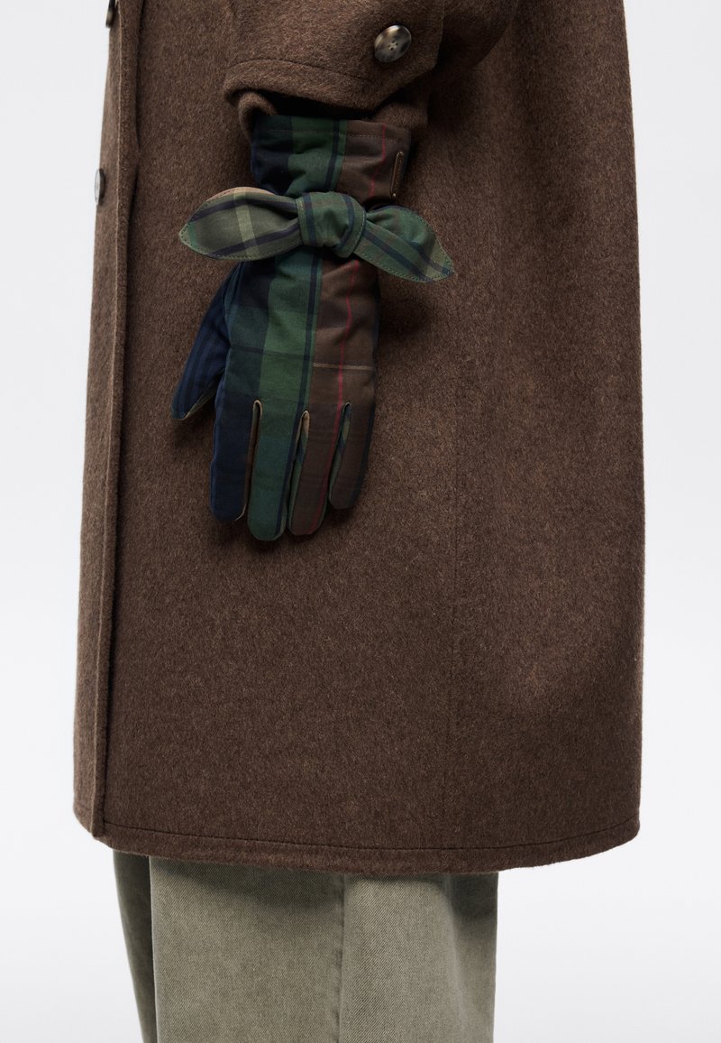 Brązowy wełniany płaszcz z zielono-niebieską kraciastą rękawiczką z detalem w postaci kokardki, prezentujący mieszankę tekstur i kolorów.
