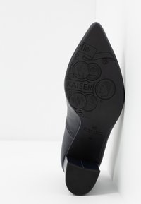 Escarpin à bout pointu en cuir bleu marine avec une semelle noire texturée présentant des motifs de pièces embossées et des détails de logo au talon.