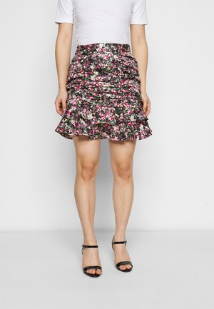Jupe mini florale avec ourlet à volants, présentant un fond noir avec des fleurs roses et blanches ; assortie à des sandales à talons noires.