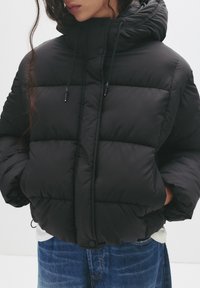 Vrouw die een zwarte gewatteerde jas met capuchon en zijzakken draagt, in combinatie met blauwe jeans, tegen een effen witte achtergrond.