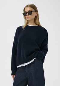 Námornícky modrý pletený sveter s voľným strihom a okrúhlym výstrihom, položený na bielom tričku. Model nosí čierne uhlové slnečné okuliare.