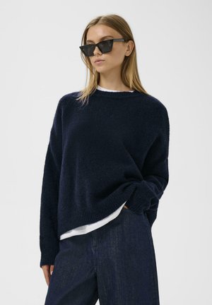 Námornícky modrý pletený sveter s voľným strihom a okrúhlym výstrihom, položený na bielom tričku. Model nosí čierne uhlové slnečné okuliare.