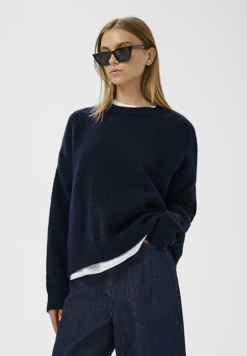 Námornícky modrý pletený sveter s voľným strihom a okrúhlym výstrihom, položený na bielom tričku. Model nosí čierne uhlové slnečné okuliare.