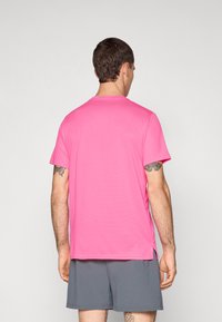 Rosa Kurzarmshirt mit Rundhalsausschnitt aus leichtem Stoff. Hat seitliche Schlitz und eine dezente Textur. Getragen mit grauen Shorts.