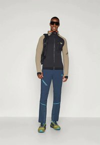 Juodas ir smėlio spalvos sportinis džemperis, tamsiai mėlynos kelnės su mėlynomis detalėmis, tvirti žali batai ir akiniai nuo saulės. Yra užtrauktukas ir logotipo detalės.