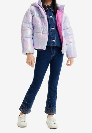 Frau trägt eine irisierende Steppjacke über einer Jeansjacke und dunkelblaue Jeans mit Strassverzierungen sowie weiße Turnschuhe.