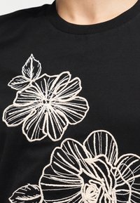 Tissu noir avec de grands motifs floraux brodés en blanc formant des pétales de fleurs et des feuilles sur un vêtement près de la zone du cou.