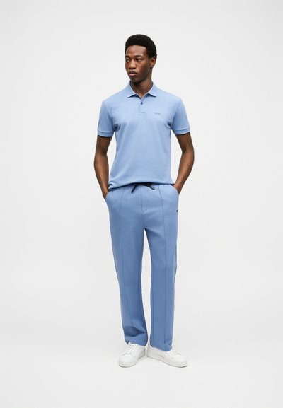 Homme debout, les mains dans les poches, portant un polo bleu clair, un pantalon assorti et des baskets blanches, sur un fond uni.