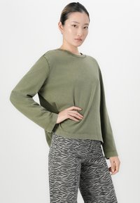 Žena v dlouhém olivově zeleném topu s jednou rukou na boku a šedými kalhotami se zebřím vzorem na jednoduchém bílém pozadí.