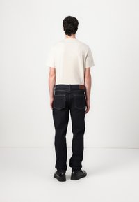 Muž stojí čelom otočený k stene, oblečený v béžovej košeli s krátkym rukávom, tmavomodrých džínsoch a čiernych topánkach na bielom pozadí.