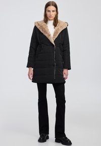 Černý vypolstrovaný kabát s béžovou umělou kožešinou na kapuci, staženým pasem a zipem. Má dlouhé rukávy a je kombinován s černými kalhotami a botami.