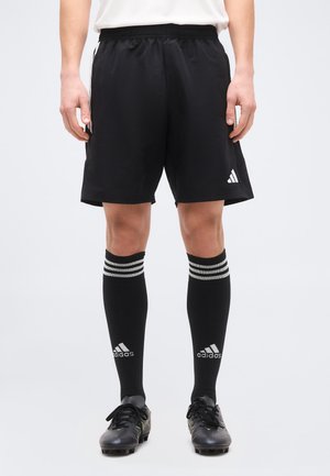 Mees, kes kannab musta värvi Adidas spordipükse, musti Adidas kolme valge triibuga põlvikuid ja musti kaitsvaid jalanõusid, seisab valge tausta ees.