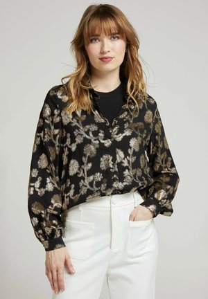 Blouse noire à manches longues avec boutons, présentant des motifs floraux métalliques en or et en argent. Manches longues avec des poignets, accompagnée de pantalons blancs taille haute.