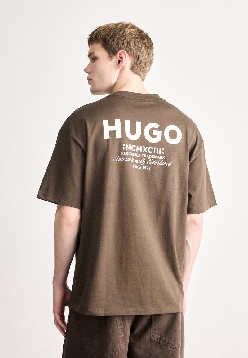 Brązowy bawełniany t-shirt z dużym białym napisem „HUGO” na plecach oraz dodatkowymi nadrukami. Krótkie rękawy, luźny krój.