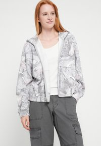 Sudadera corta con capucha en gris claro con un sutil patrón de camuflaje en rosa y gris, con cierre de cremallera y tejido suave. Combinada con pantalones cargo grises.