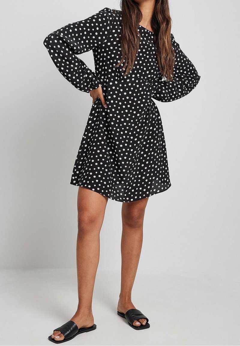 Robe noire à pois avec des manches longues, coupe ample et col rond. Portée avec des chaussons plats noirs. Texture de tissu lisse.