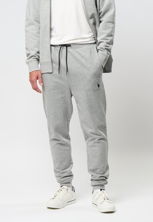 Mand iført lysegrå joggingbukser med sort snor, matchende grå hættetrøje med lynlås, hvid t-shirt og hvide sneakers, stående med hænderne i lommerne.