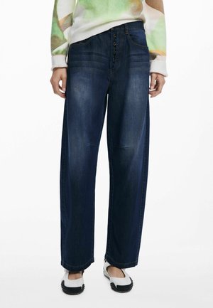 Donkerblauwe wijde jeans met een knoopslag, vijf zakken en een lichte vervaging langs de benen. Gepaard met witte instappers.