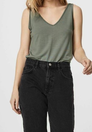 Kvinde iført ærmeløs olivengrøn V-hals top indstukket i høj talje sort jeans mod en ensfarvet lys baggrund.