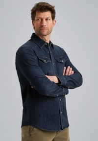 Ciemnoniebieska koszula dżinsowa z zapięciem na guziki z przodu, spiczastym kołnierzykiem i kieszeniami na piersi. Posiada długie rękawy i dopasowany krój. Gładka tekstura.