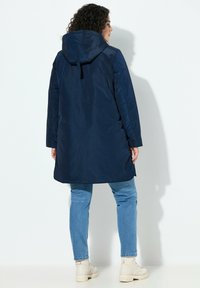 Tummansininen, hupullinen parkatakki, joka on valmistettu sileästä, vettähylkivästä kankaasta. Siinä on rento istuvuus ja sivutaskut, yhdistetty vaalean sinisten farkkujen ja beigen väristen nilkkureiden kanssa.