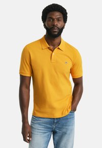 Gelbes Polohemd mit klassischem Kragen und Drei-Knopf-Leiste, aus strukturiertem Baumwollstoff, getragen mit hellblauen Jeans.