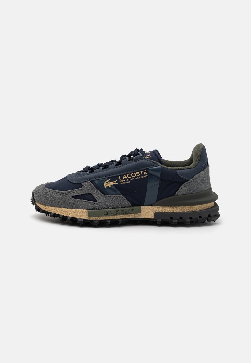 Sneakers Lacoste con tomaia blu navy, dettagli in suede grigi e suola in gomma testurizzata. Il marchio dorato sul lato aggiunge un tocco di dettaglio.