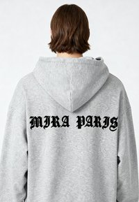 Felpa grigia con un grande logo nero "MIRA PARIS" sul retro, caratterizzata da una vestibilità rilassata e un cappuccio ampio. Tessuto morbido e accogliente.