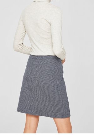 Femme portant une jupe à carreaux gris et noir longueur genoux et un haut à manches longues beige clair, debout dos à la caméra.