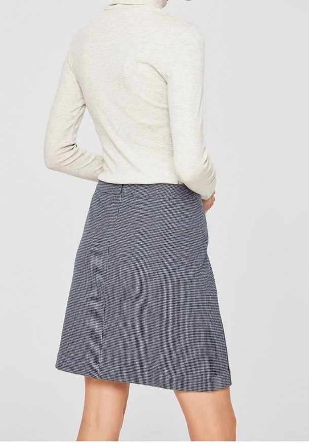 Femme portant une jupe à carreaux gris et noir longueur genoux et un haut à manches longues beige clair, debout dos à la caméra.