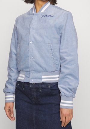 Kobieta ubrana w jasnoniebieską kurtkę varsity z sztruksu z białymi paskowanymi ściągaczami przy mankietach i pasku, w połączeniu z ciemną jeansową spódnicą.