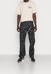 Camiseta de algodón beige con un logotipo en el bolsillo del pecho, combinada con pantalones negros con un patrón de líneas blancas. Estilizada con zapatillas negras.