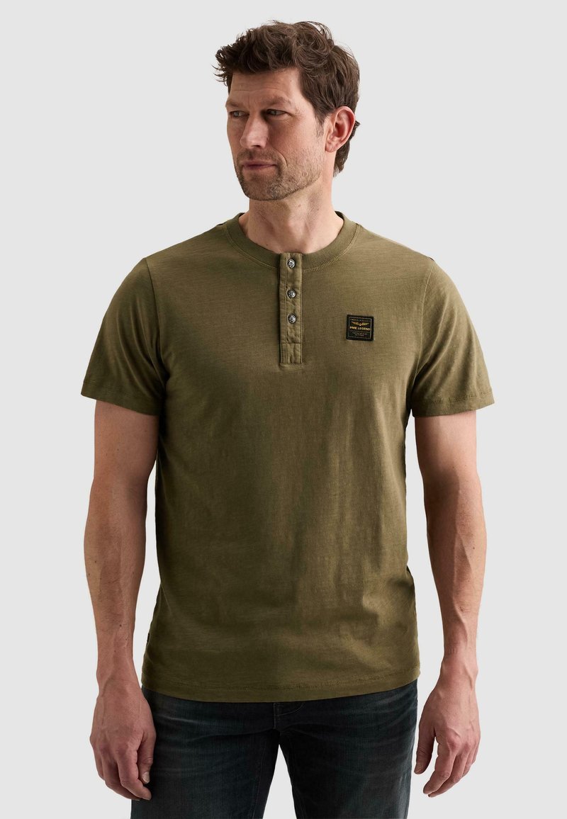 Homme portant un t-shirt henley à manches courtes vert olive avec trois boutons et un petit patch noir sur la poitrine, debout sur un fond uni.